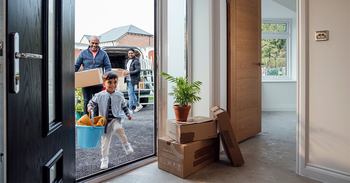 A family of three arrives at their new home. Their child is carrying a bucket into the front door, and their full service moving company has already dropped some moving boxes off in their new home.