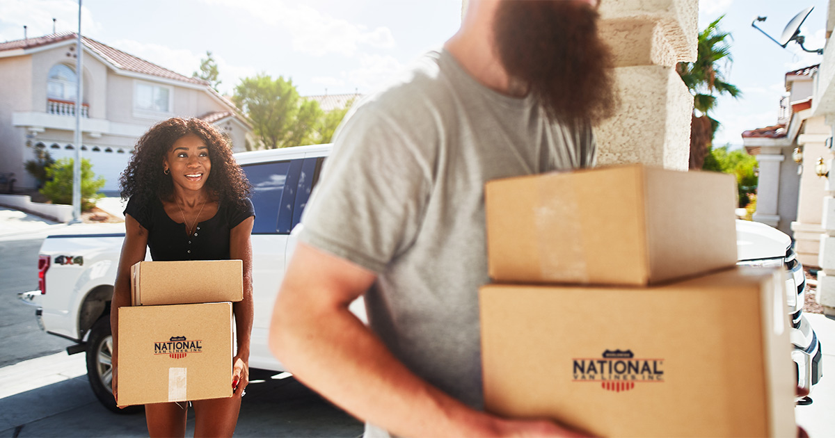 A young couple is walking from a white truck parked on the driveway of their new home. They are carrying National Van Lines moving boxes. They've just moved long distance, and started planning a move step by step when they initially found out. 