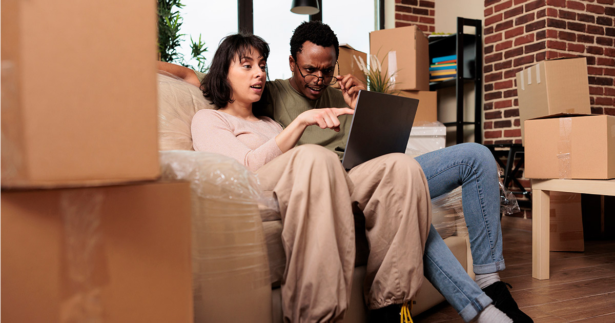 A couple sits together on a couch. They are surrounded by moving boxes. They are listing a home for sale, and are starting on their move planning.