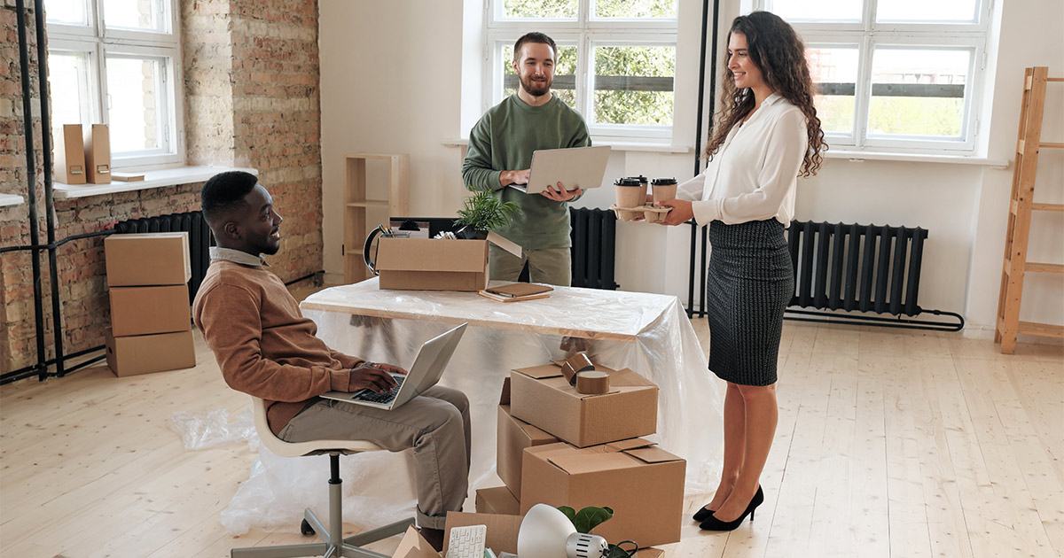 Three employees discuss the upcoming corporate move for their company. They are surrounded by moving boxes and packing supplies. They will be working with a corporate moving company. 