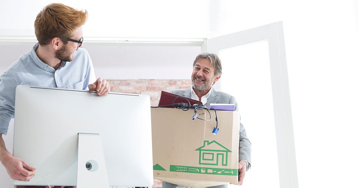 After a long distance corporate move, two employees greet each other in their new office. The man on the right holds a moving box full of desk supplies, with the man on the left plugs in his computer. 