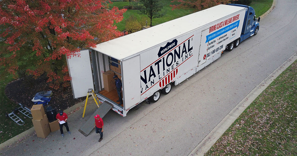 A National Van Line moving truck sits in a residential area. Two National Van Lines employees review a checklist and stand near the loading ramp. This truck is being used for long distance moves. 