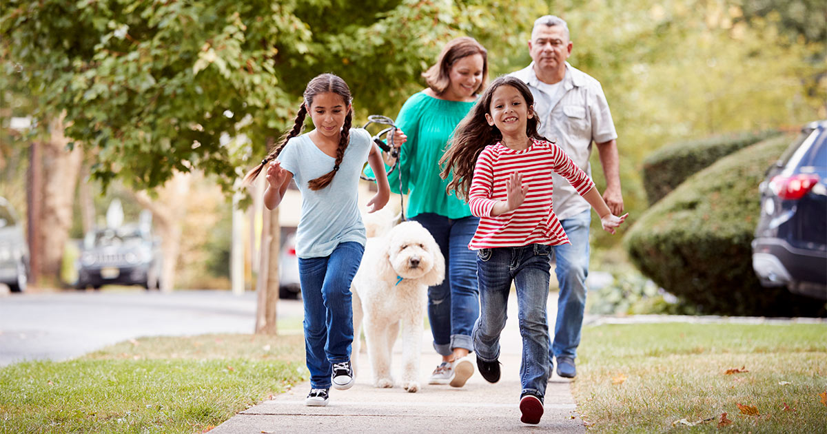 A family of four has just moved to their new neighborhood after a long distance move, and are taking their large white poodle mix dog on a walk. The children run happily in the forefront.
