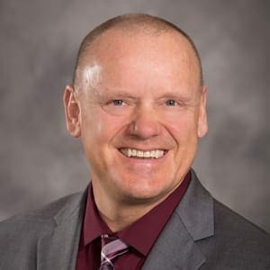 A person with short hair is smiling broadly. They are wearing a maroon dress shirt, a striped tie, and a gray suit jacket. The background is blurred gray tones.