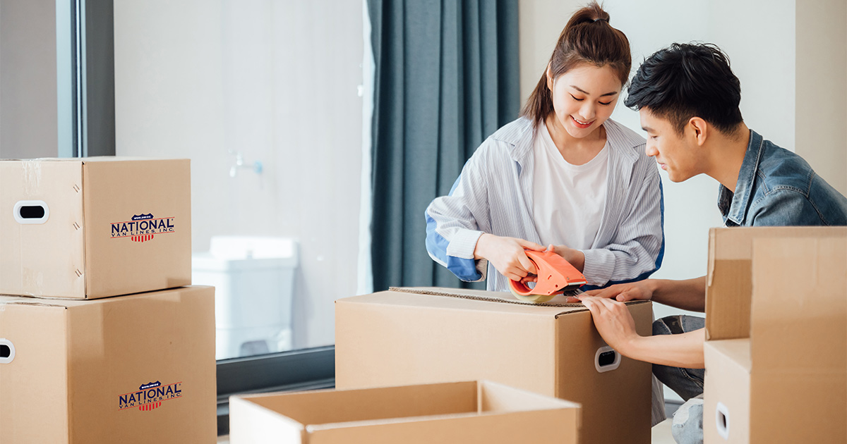 A young couple pack National Van Lines moving boxes in their living room. They are preparing for a long distance move. They will be using secure storage along the way to store items during the interim.