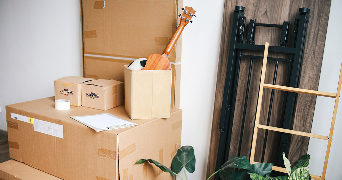 National Van Lines moving boxes are stacked in a room, with a table, ladder, and planet nearby. There is a box with a ukulele and soccer ball. There are also packing supplies and a checklist. These boxes are being prepared for a long distance move, and will be put into secure storage along the way.