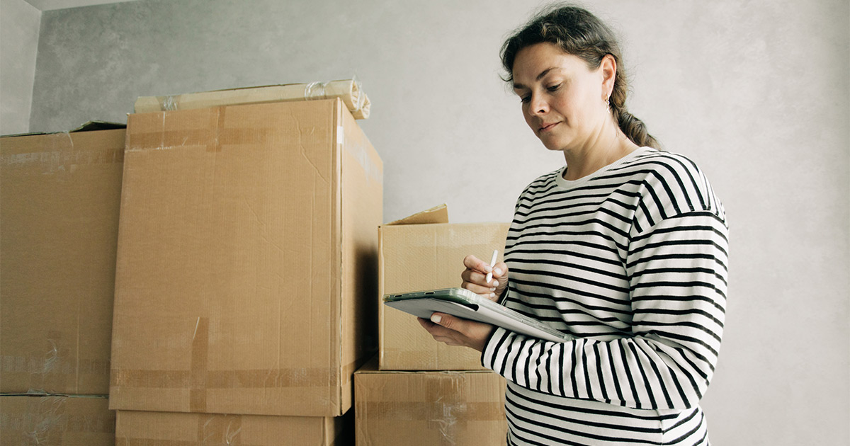 A woman who is diagnosed with ADHD stands near some stacks of moving boxes. She is using a checklist to making moving with ADHD more organized and manageable. 