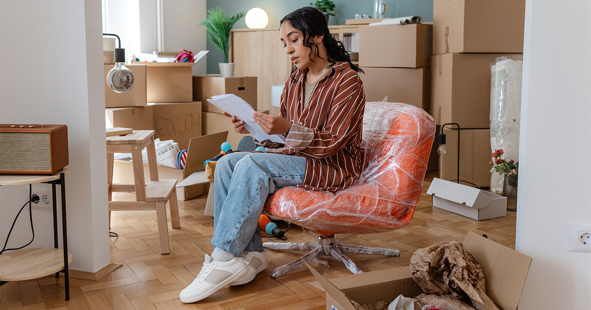 A woman sits on a wrapped chair amongst many moving boxes. She is reviewing her moving valuation paperwork before a long distance move.