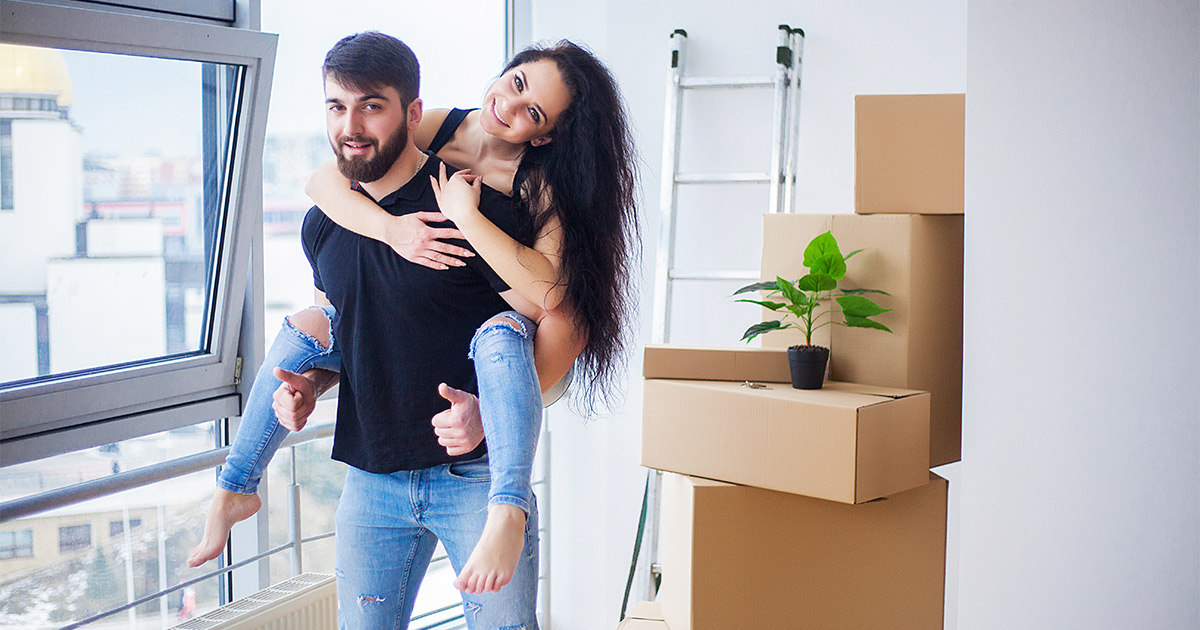 A young couple stands near their apartment window. The man is carrying the woman on his back and they are giving a smile. There are moving boxes nearby. They researched best moving companies for their long distance move, and were able to get a moving company that made their apartment move extremely easy. 