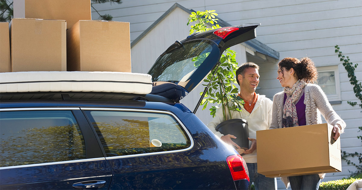A couple unloads their car while smiling. They are outside their new home and are carrying moving boxes. They researched best moving companies prior to moving and had an excellent experience with the moving company they chose. 