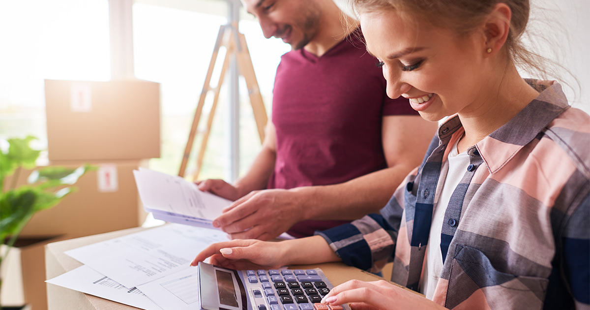 A couple, smiling, has laid out paperwork on top of stacked moving boxes. They are moving cross country, and they started by finding out the answer to the question, "What does it cost to move cross country?" 