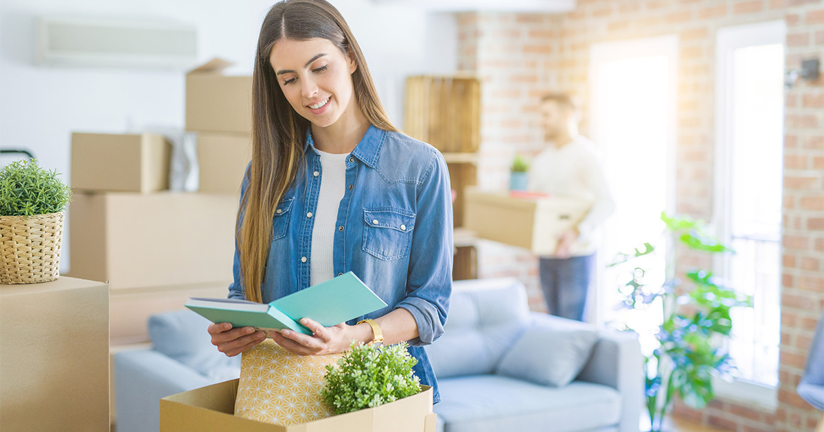 A woman holds a notebook over an open moving box. A man stands in the background holding a moving box with a plant balanced on top. There are moving boxes surrounding them everywhere in their new living room. The woman is smiling because the move went well, and she went in knowing how much it costs to move cross country. 