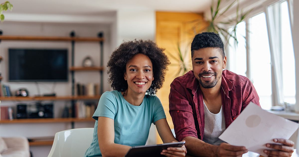 A couple looks over their finances and budget at their kitchen table. They are smiling. The man holds a piece of paper, and the woman holds a tablet. They are getting ready to move cross country, and are looking into the costs. 