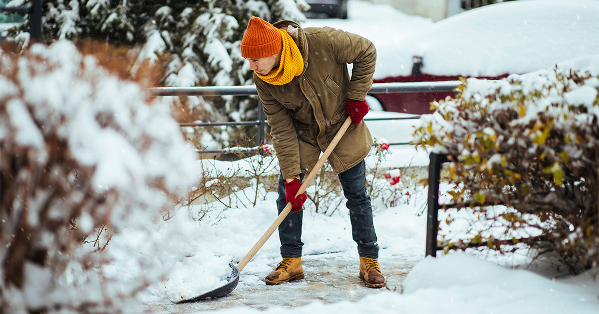 A man in a hat, gloves, scarf and a coat shovels his front walkway in preparation for a moving crew. 