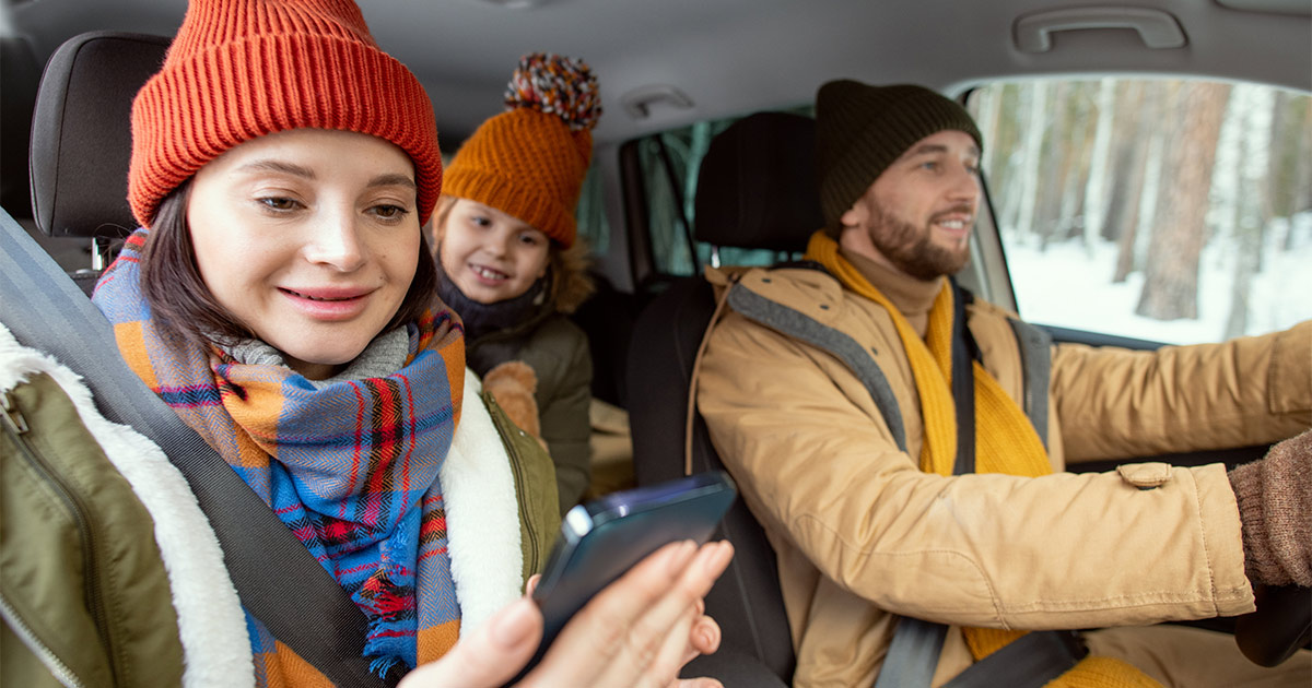 Two parents and a child drive in a snowy environment. The mother smiles while checking her smartphone, as she has just received a positive update on her long distance move. 