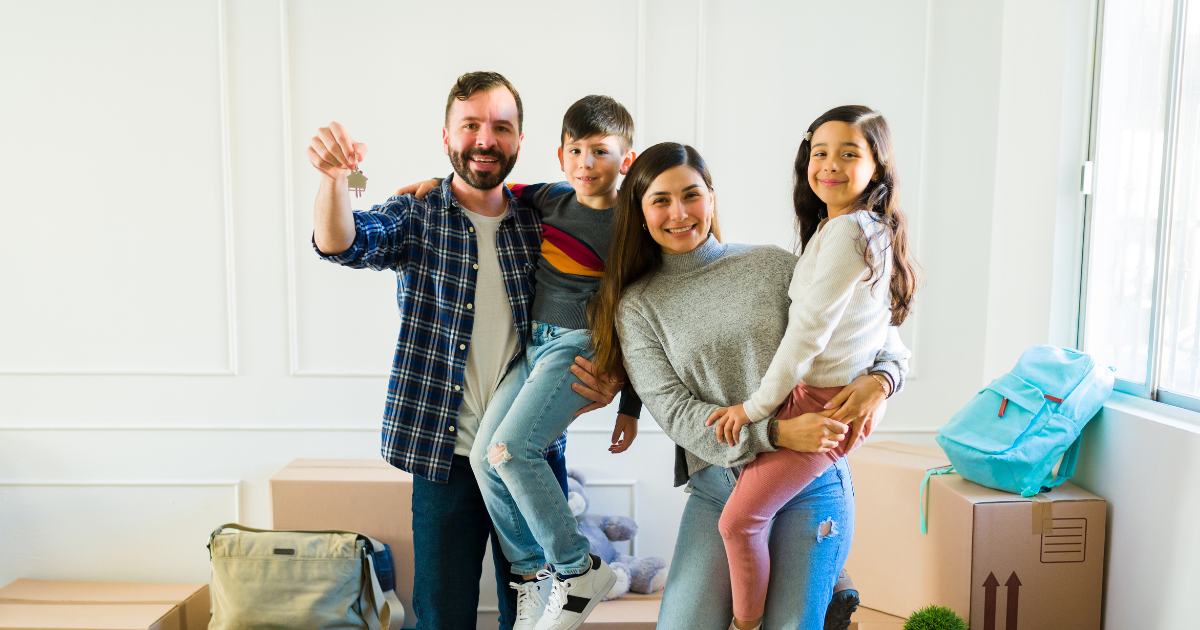 A family of four stands amongst moving boxes and packing supplies. The father holds keys to their new home after they completed a move abroad.