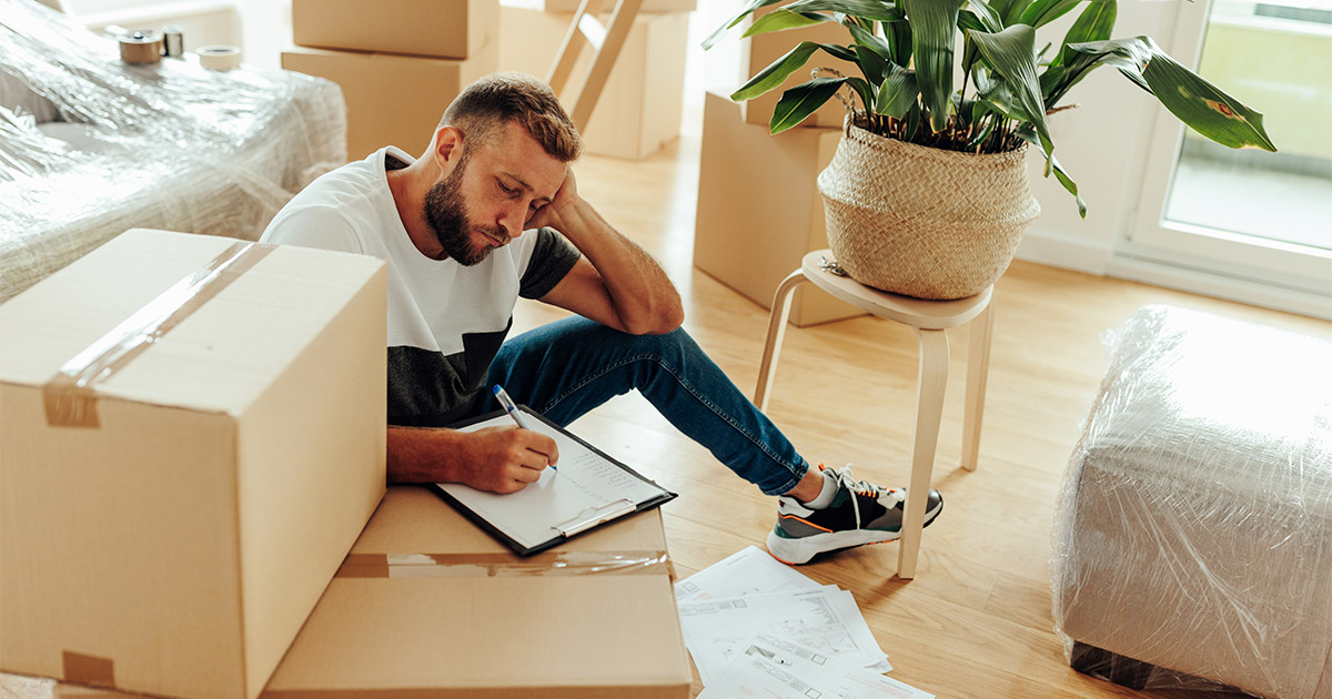 A man sits amongst moving boxes and packing supplies, and writes on a clipboard. The clipboard lists what movers won't move. He is preparing for an upcoming long distance move.