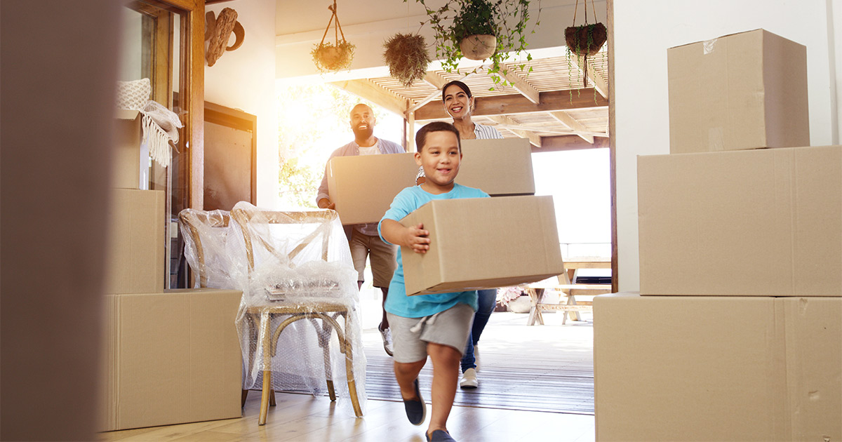 A family of three carries moving boxes happily into their new home. The long distance movers have just arrived and delivered their items and furniture. 