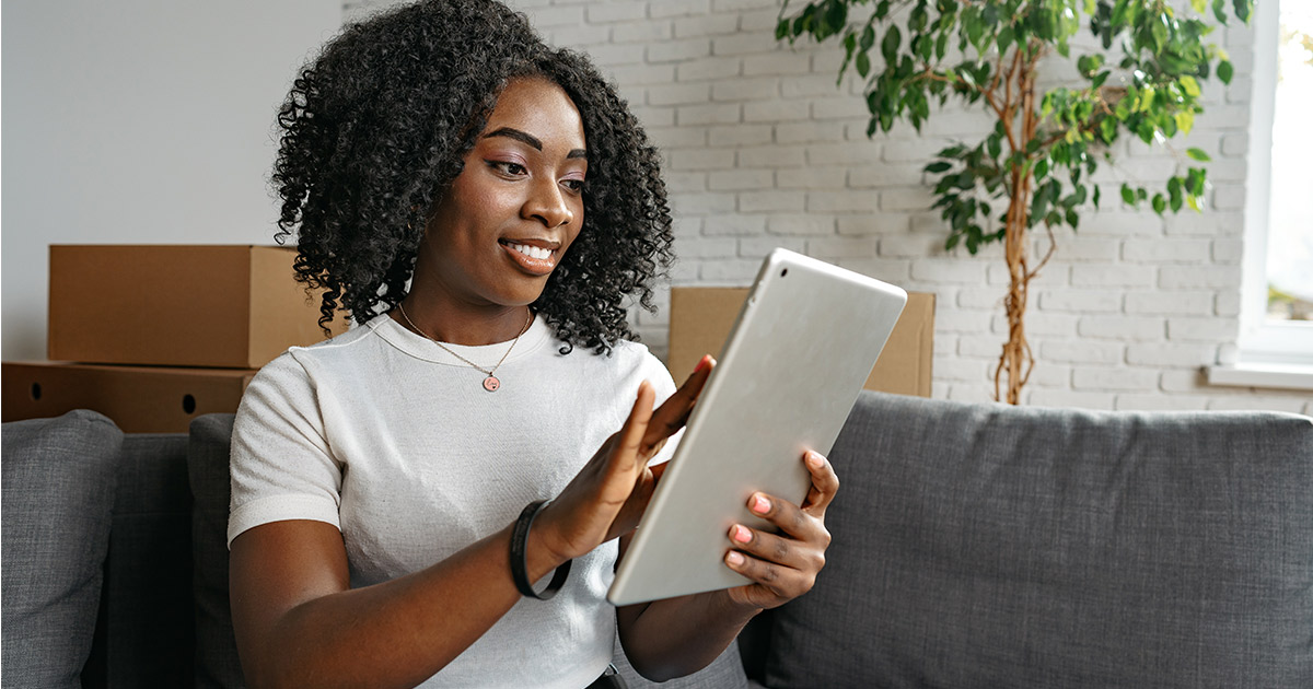 A woman, smiling, sits on her couch and looks at her tablet. Moving boxes are stacked behind the couch. She is using the term "long distance moving companies near me" in different search engines and apps to get an idea of what moving companies she can choose for her upcoming interstate move.