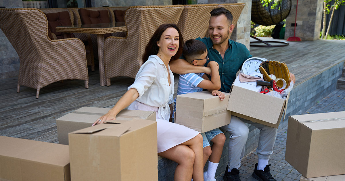 A family of three sits on their new deck of their new house after a long distance move. They are smiling. The father is hugging the son. They are surrounded by moving boxes. One of the tactics they used during the planning process was "long distance moving companies near me" to understand their options.