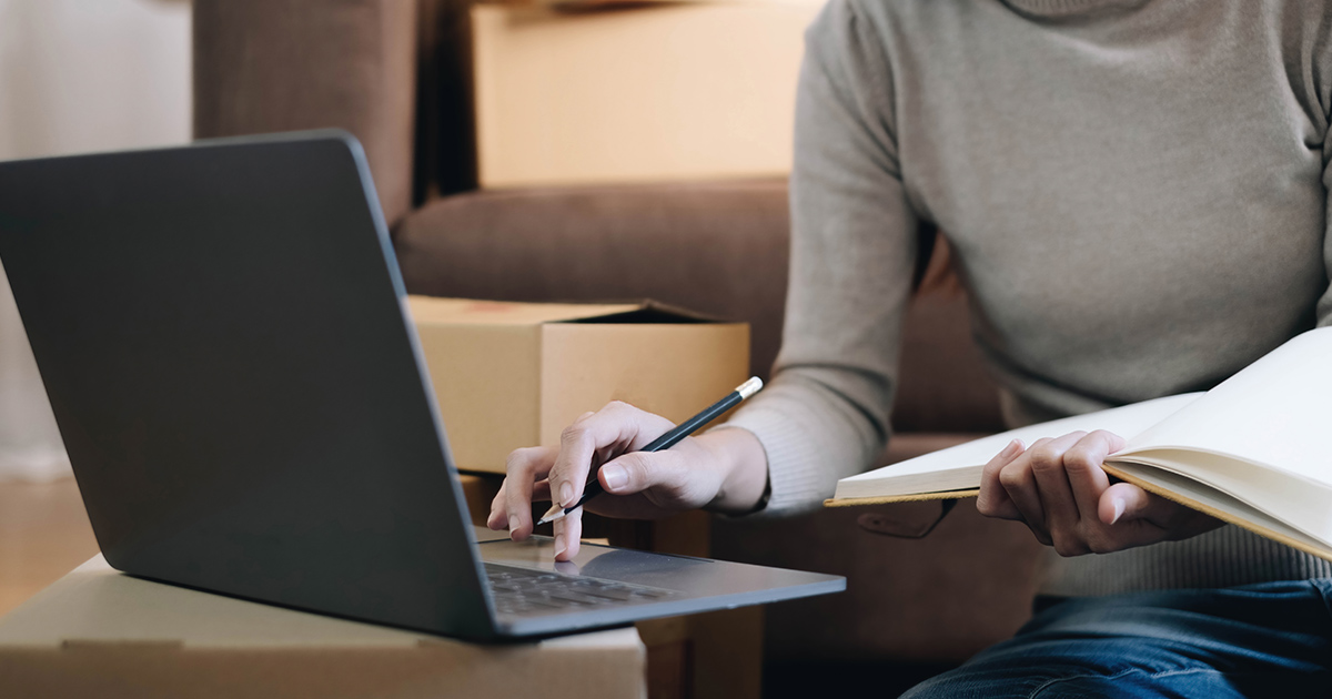 A laptop computer sits on top of a moving box. Someone sits in front of it while holding a notebook and pencil. They are using the term "long distance moving companies near me" in search engines to research long distance moving companies.