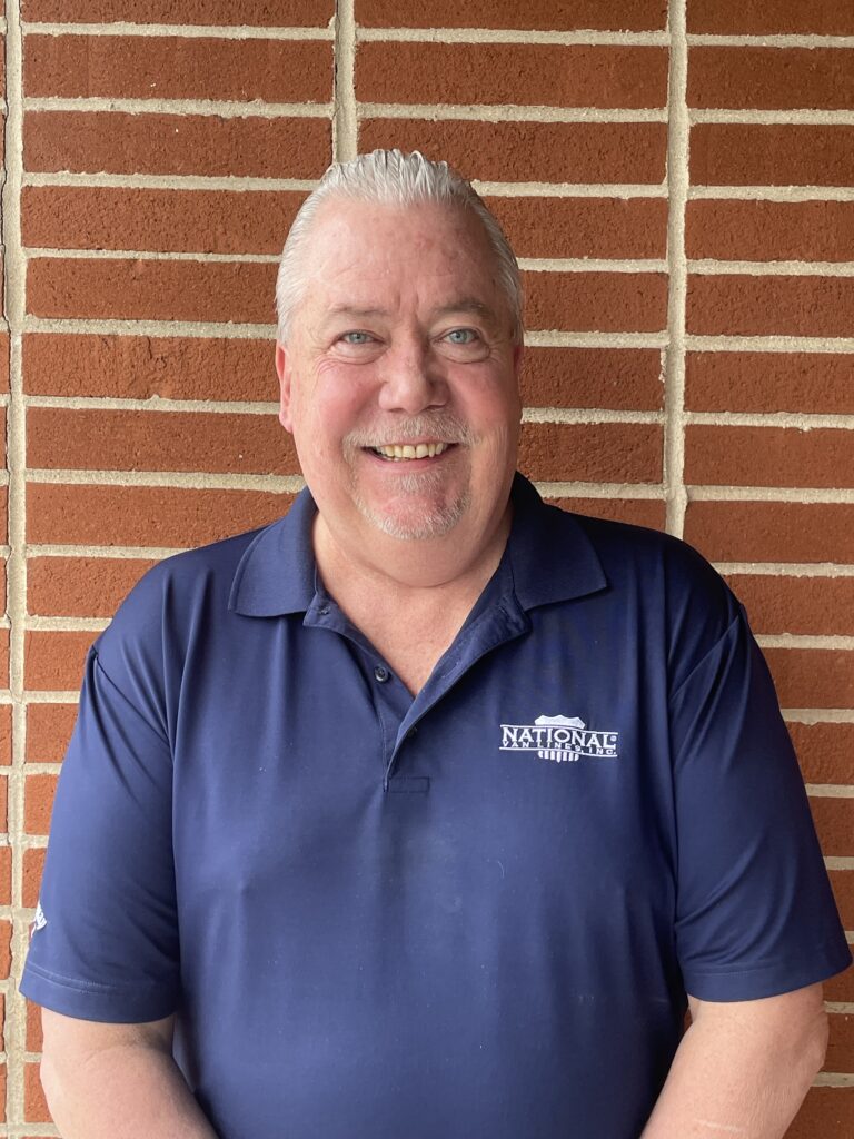 A smiling man with short white hair stands in front of a brick wall. He is wearing a navy blue polo shirt with a 