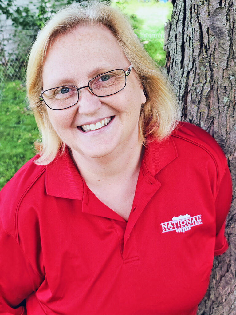 A smiling person with light hair and glasses leans against a tree trunk. They are wearing a red polo shirt with a 