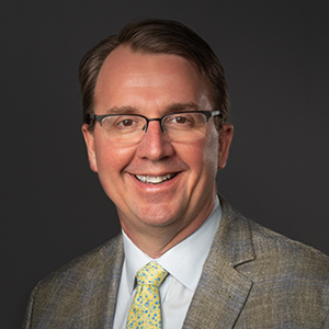 A man with short brown hair and glasses is smiling at the camera. He is wearing a gray suit jacket, a white shirt, and a yellow patterned tie. The background is solid dark gray.