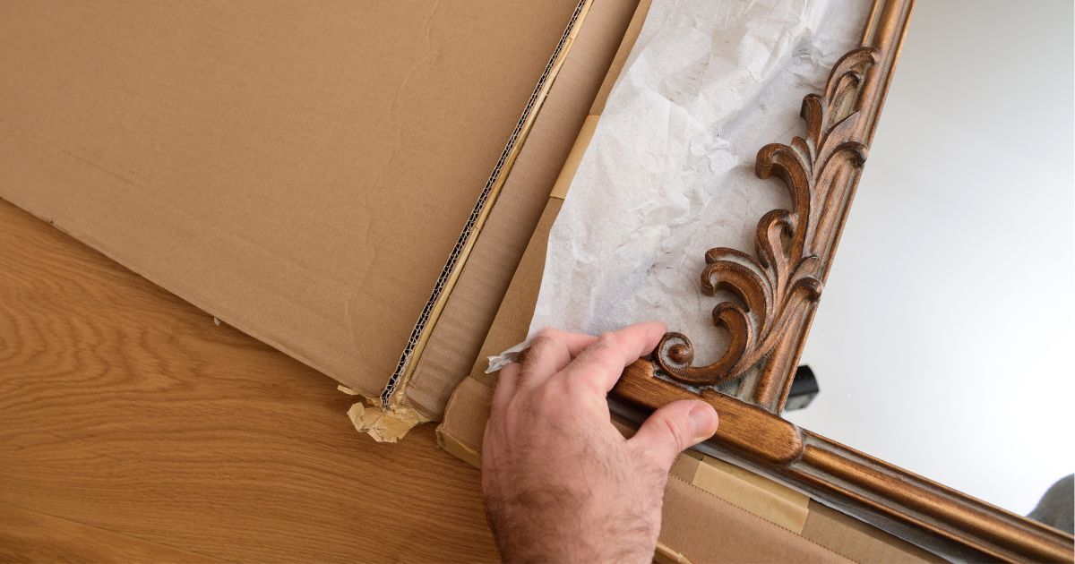 A person carefully places an ornate mirror into a moving box for a long distance move.
