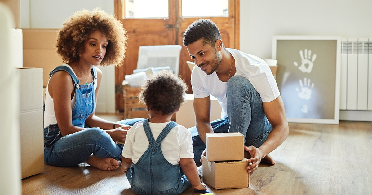 Two parents sit with their young child on the floor of their home amongst moving boxes and packing supplies.