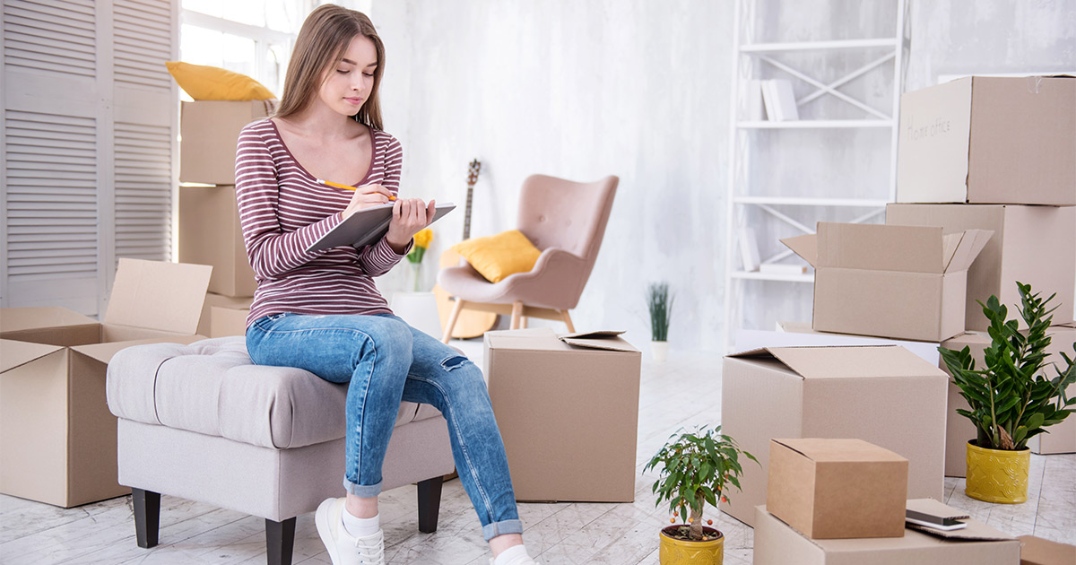 A woman makes a list in her notebook for her long distance move while sitting amongst moving boxes and packing supplies.