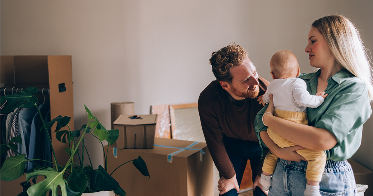 A young couple hold their baby while smiling. They are standing amongst moving boxes and packing supplies and are preparing for a long distance move. 