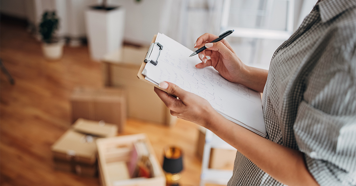 A person takes notes on a checklist in preparation for an upcoming long distance move. This person is standing amongst moving boxes and packing supplies. 