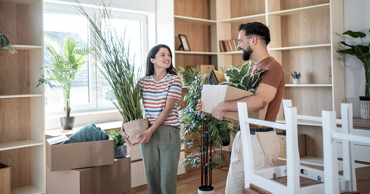 A man and woman have a cheerful conversation while they carry items from their bookshelf. They are preparing for a long distance move and are wondering how long it will take to move long distance.