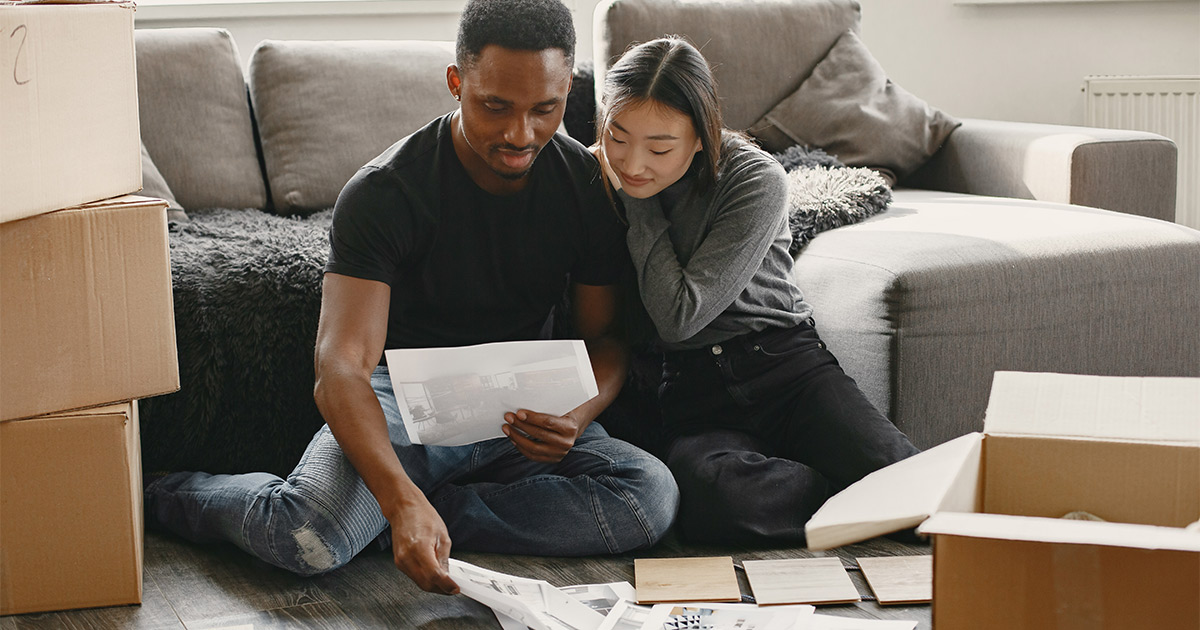 A young couple sits in their living room amongst moving boxes and packing supplies. They are considering what it would be like to move cross country, and are exploring their options. 