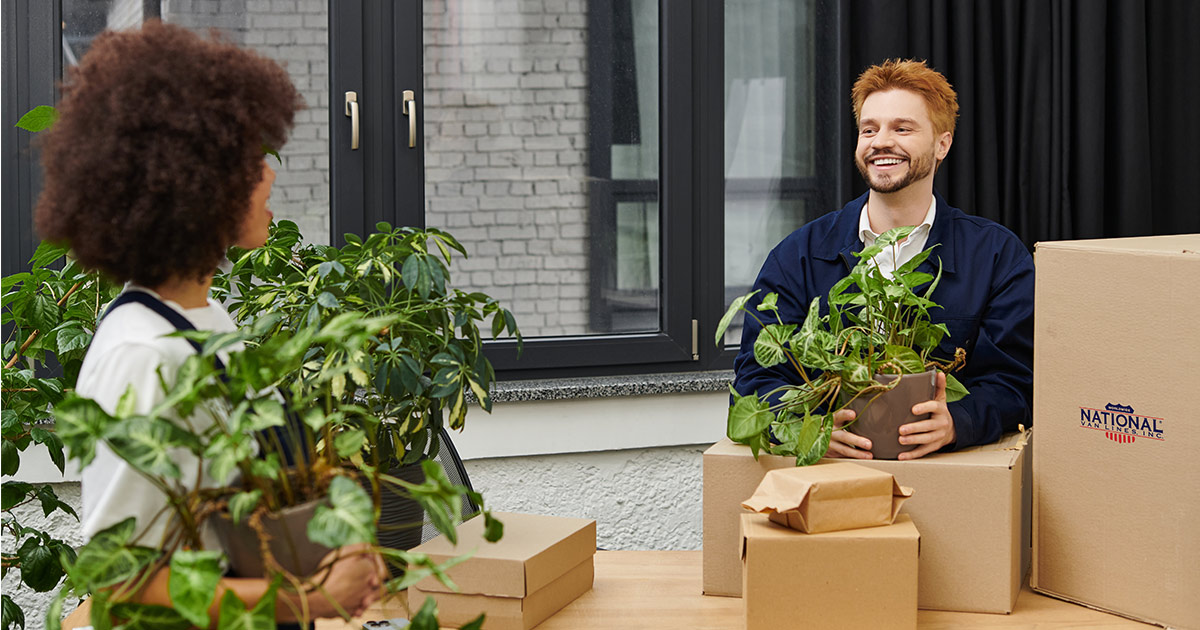 Two young coworkers pack up their desks. They are holding plants while smiling and talking to each other in an office. They are preparing for a commercial move. Their company has picked a new office. 