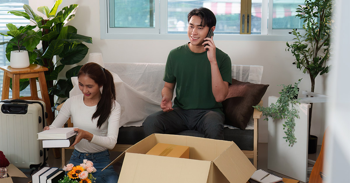A young couple sits in their living room. They are surrounded by packing supplies and moving boxes. The woman stacks books and prepares to put them in an open moving box. The man is on the phone with a moving company doing research. He wants to avoid common moving mistakes.