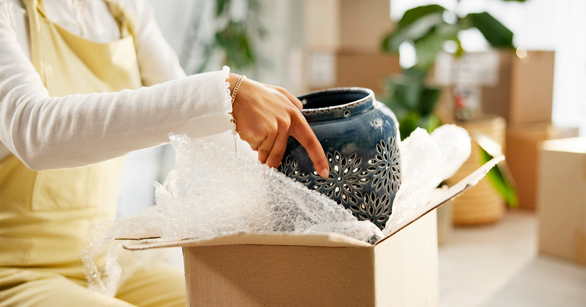 A woman packs a fragile vase into bubble wrap and a moving box. She is preparing for a specialty move. 
