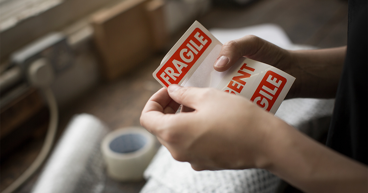 Someone holds a fragile sticker label in their hands. In the background are packing supplies. This person is preparing for a specialty move, cross country. 