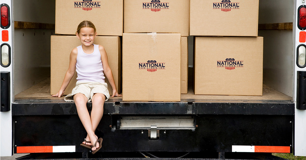 A young girl sits in the back of an open moving truck, with National Van Lines moving boxes stacked behind her. She is smiling. Her parents are using National Van Lines for their long distance move, which is also going to be considered a specialty move. 
