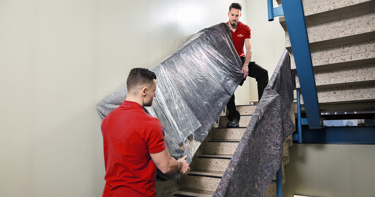 Two professional movers carry a wrapped couch down an apartment stairwell. 