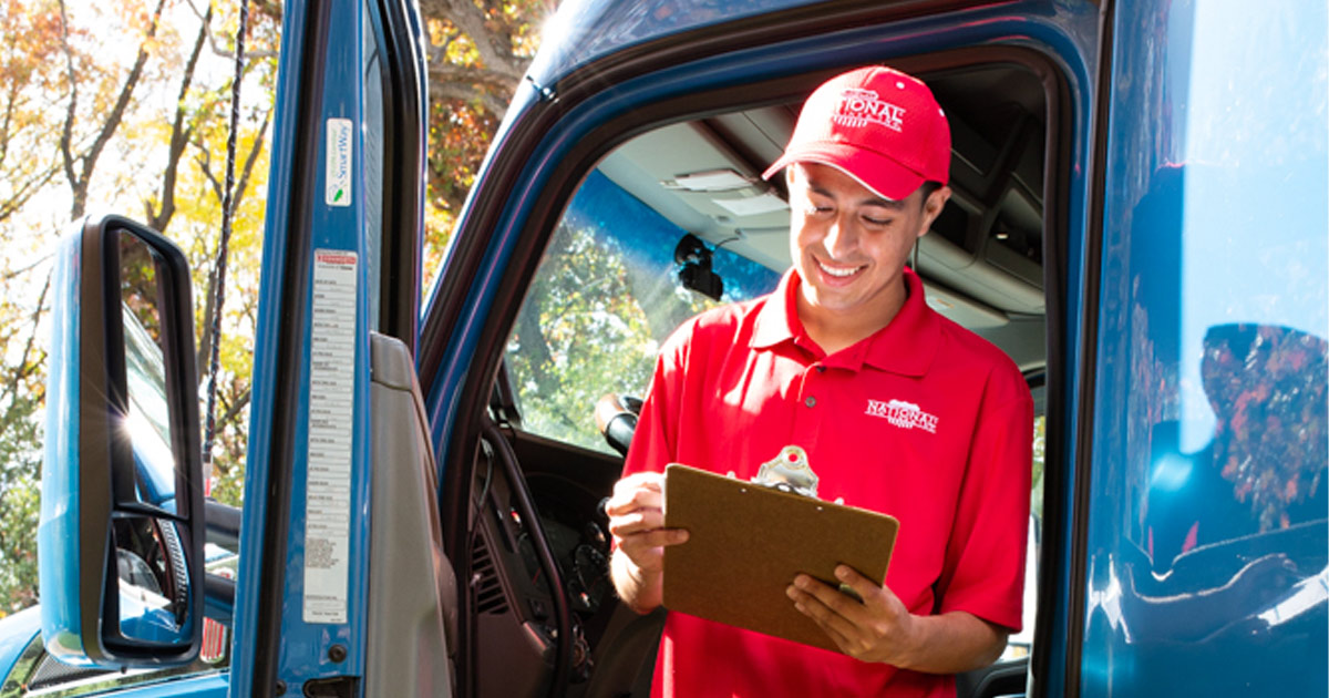 A National Van Lines driver leans against his truck and reviews a clipboard while smiling. 