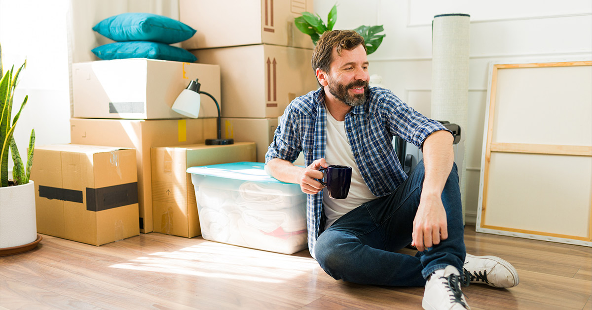 A man sits against a stack of moving boxes while holding a coffee cup and smiling. 