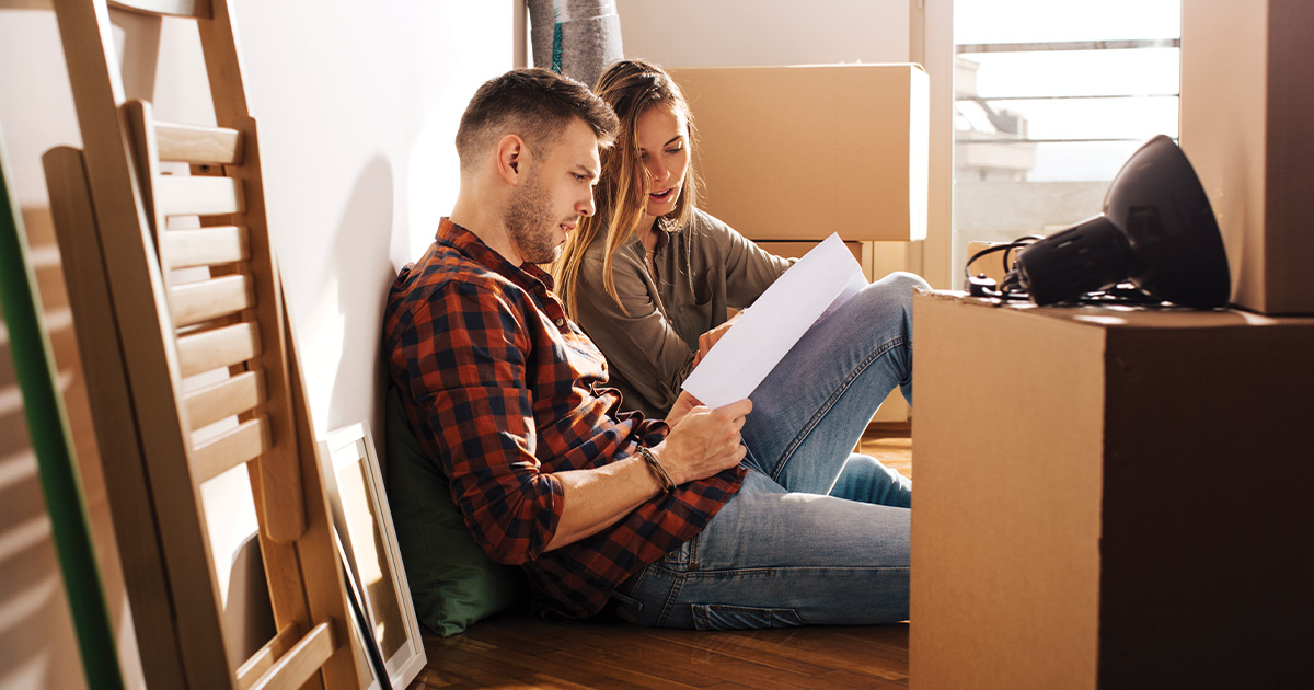 A couple sits amongst boxes and furniture, reading a document in preparation for moving day. 