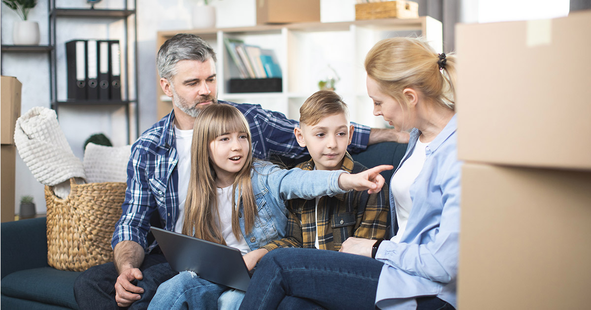 A family of four sits on their couch. The daughter has a tablet on her lap and is pointing towards some moving boxes next to her mother. The family is discussing moving cross country, and are figuring out exactly where they want to start planning. 