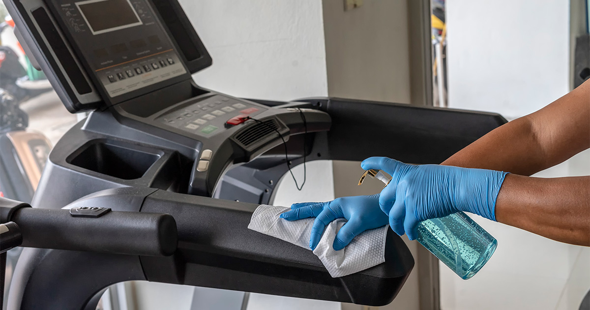 A person with gloved hands cleans the top of a treadmill. 