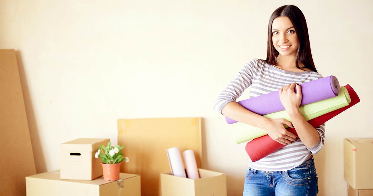 A smiling woman holds three yoga mats while surrounded by moving boxes. 