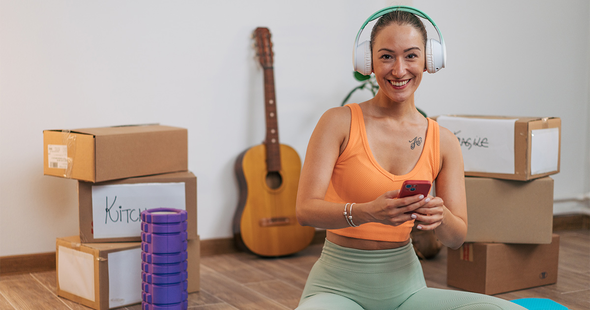 A smiling woman in fitness clothes sits amongst moving boxes with headphones on. 