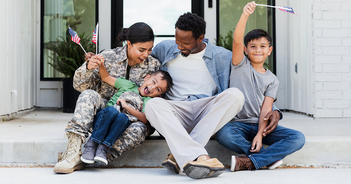 A military family sits on their front step, waving American flags. The mother is wearing an Army uniform. 
