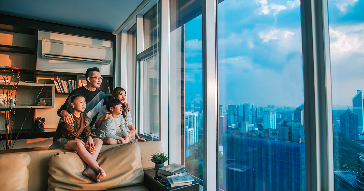 A family of four sits in the living room of their high rise apartment and looks out over a city view at sunset.
